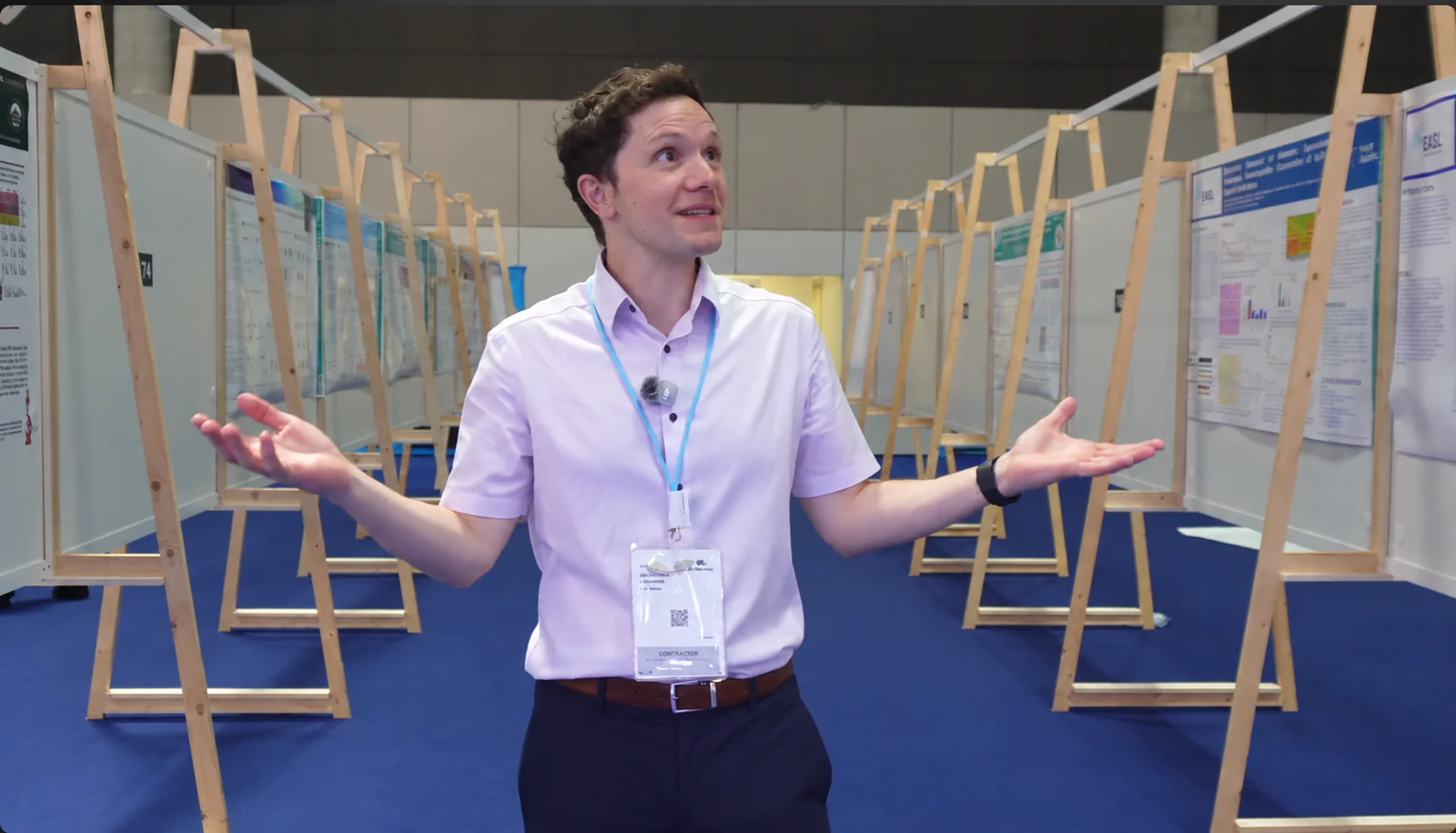 Mike Morrison in a conference poster hall, shrugging between rows of text-dense posters on wooden easels.